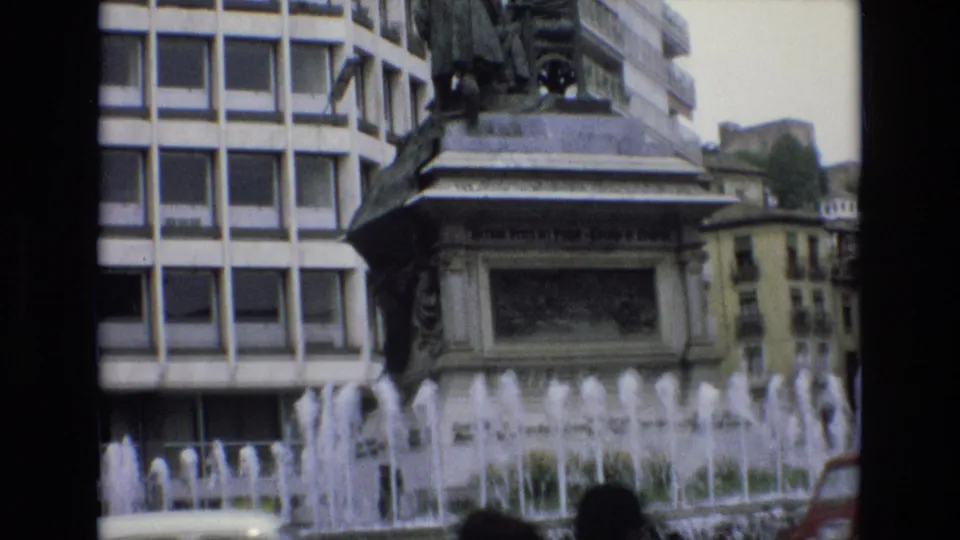 1972 Spain 8mm frame showing a city fountain before an ornate monument base, with a grid-window building and passing street traffic in the foreground.