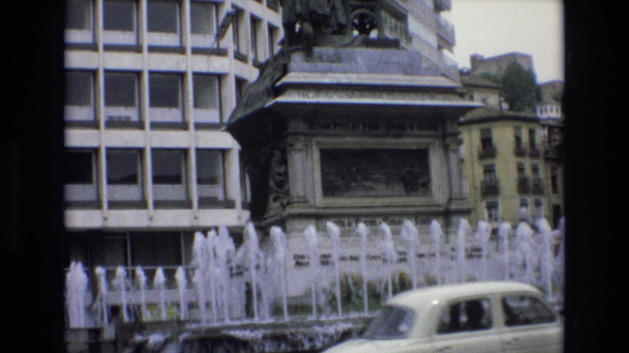 A pale vintage car passes in front of a fountain, monument base, and city buildings.