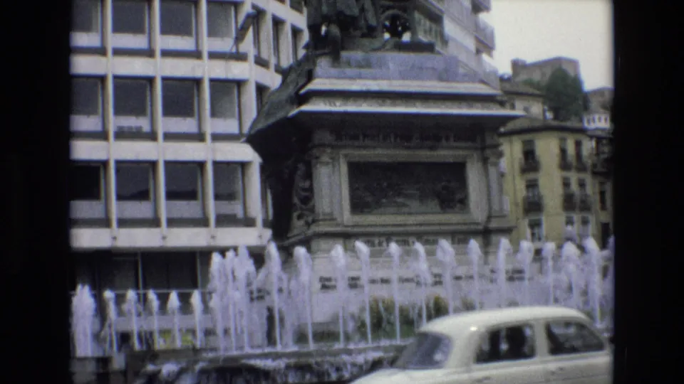 1972 Spain 8mm frame with a pale classic car crossing the foreground below a public fountain and ornate monument in an urban square.