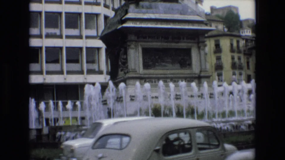 1972 Spain 8mm frame showing a rounded white car in the foreground with fountain spray, an ornate monument base, and urban buildings behind it.