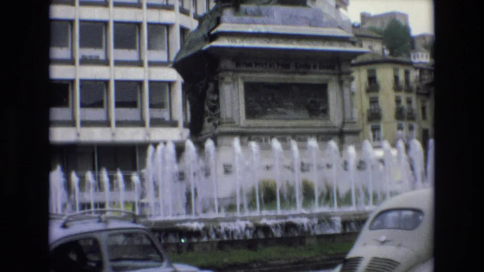 1972 Spain 8mm frame holding on the fountain and monument in a city square, with classic cars partly visible along the foreground roadway.