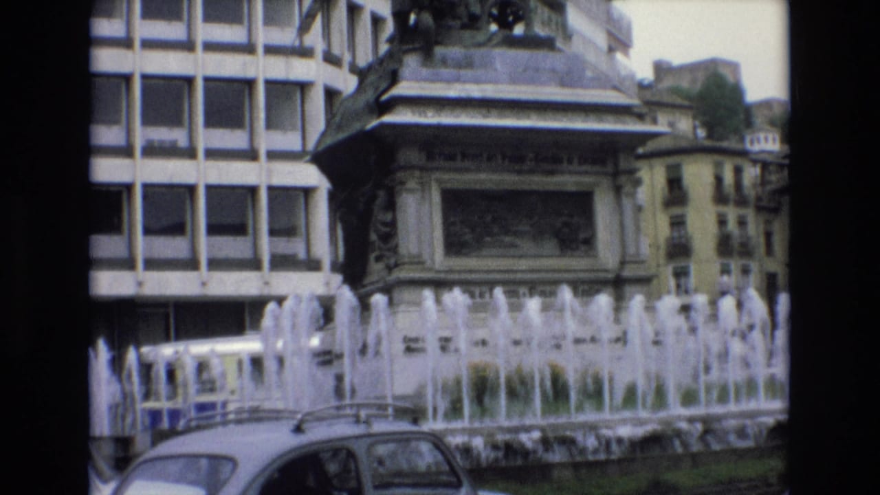 1972 Spain 8mm frame showing fountain jets, a stone monument pedestal, a modern grid-window building, older facades, and the roofline of a car at street level.