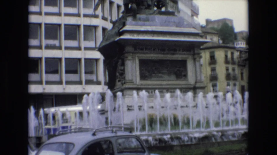 1972 Spain 8mm frame showing fountain jets, a stone monument pedestal, a modern grid-window building, older facades, and the roofline of a car at street level.