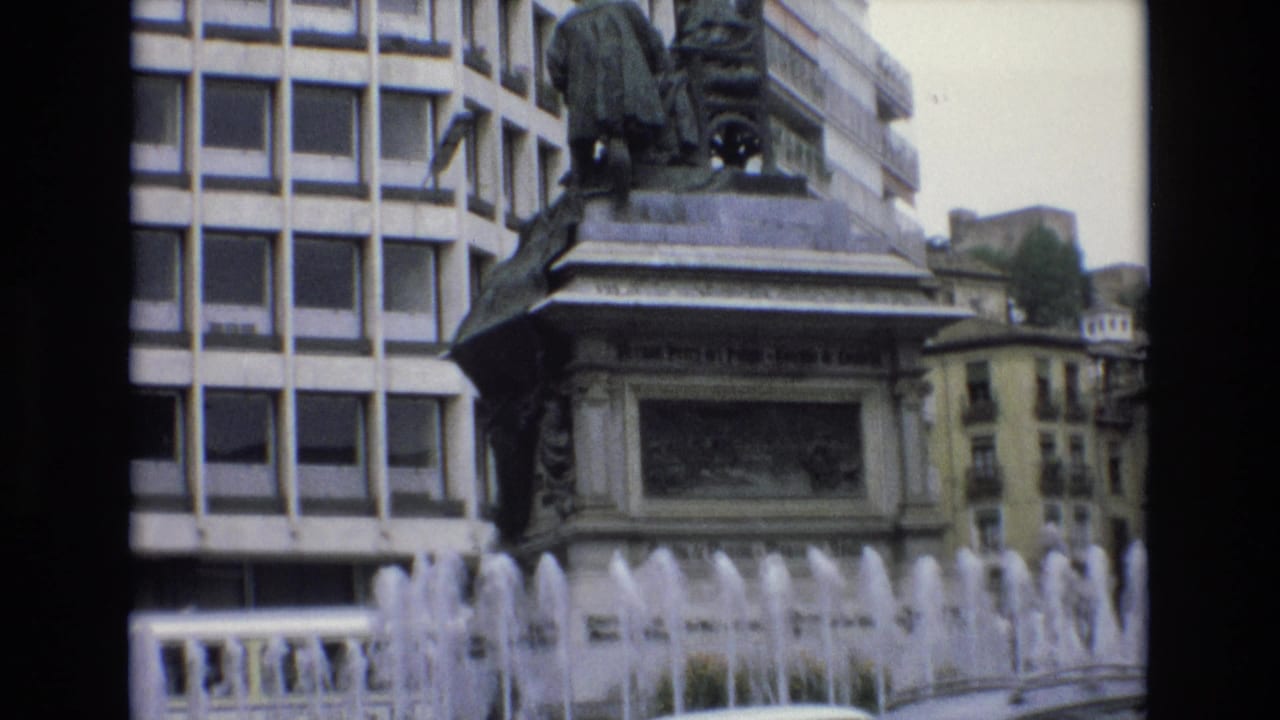 1972 Spain 8mm frame with the fountain crossing the foreground, a large sculptural monument on a stone pedestal, a modern facade behind it, and older buildings beyond.