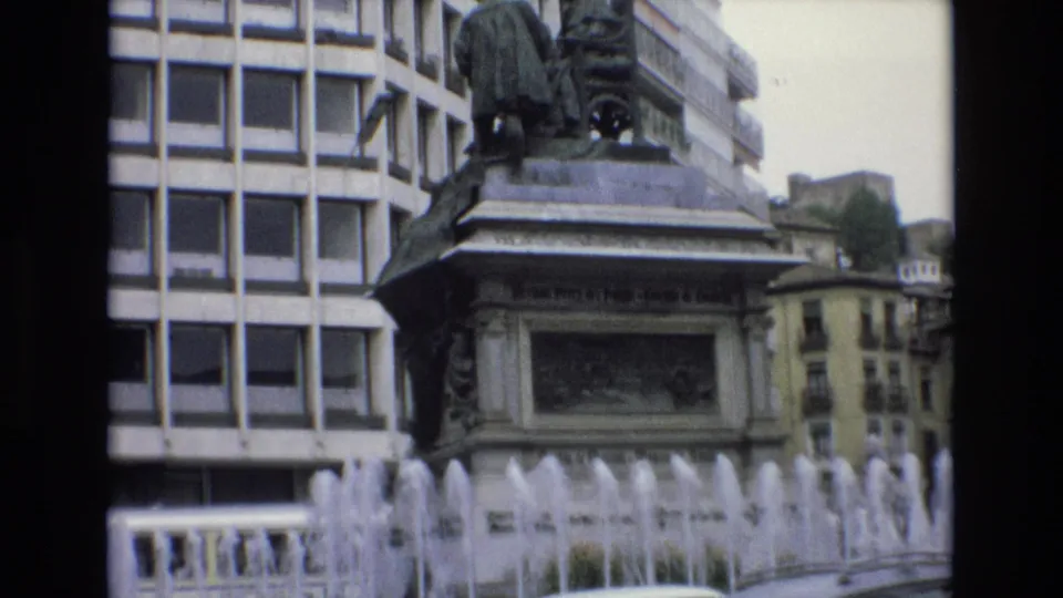 1972 Spain 8mm frame with the fountain crossing the foreground, a large sculptural monument on a stone pedestal, a modern facade behind it, and older buildings beyond.