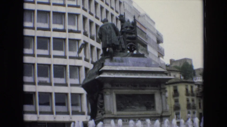 1972 Spain 8mm frame centered on the monument pedestal and sculptural figures, with a gridded modern building at left and older city architecture in the background.