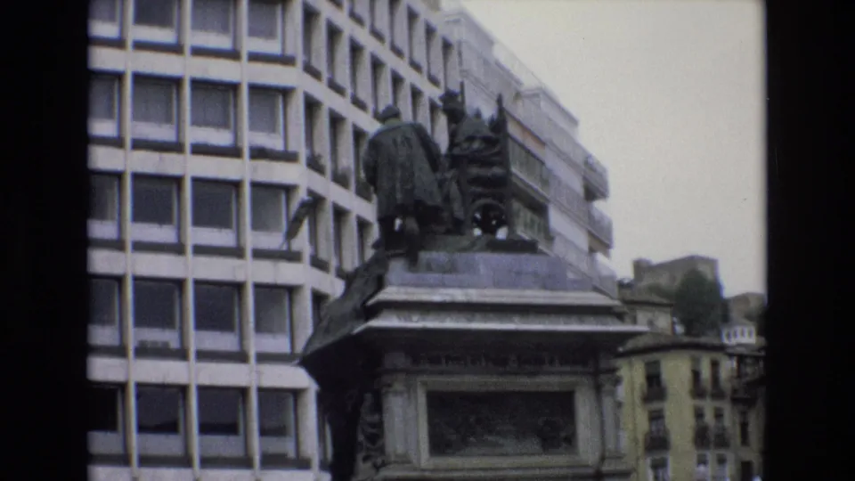 1972 Spain 8mm frame showing the statue and stone pedestal above fountain spray, framed by a modern grid-window building and older urban facades in the distance.
