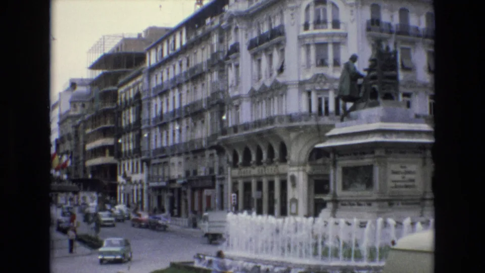1972 Spain 8mm frame showing a busy city street beside an ornate white building, with a stone fountain spraying in the foreground and traffic moving past a statue.