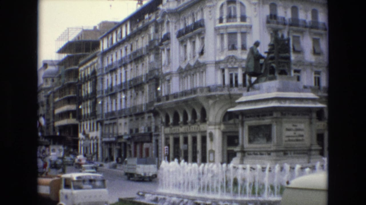 1972 Spain street view with a white van, several cars, fountain spray, a pedestal statue, and a large white building with balconies and arched windows.