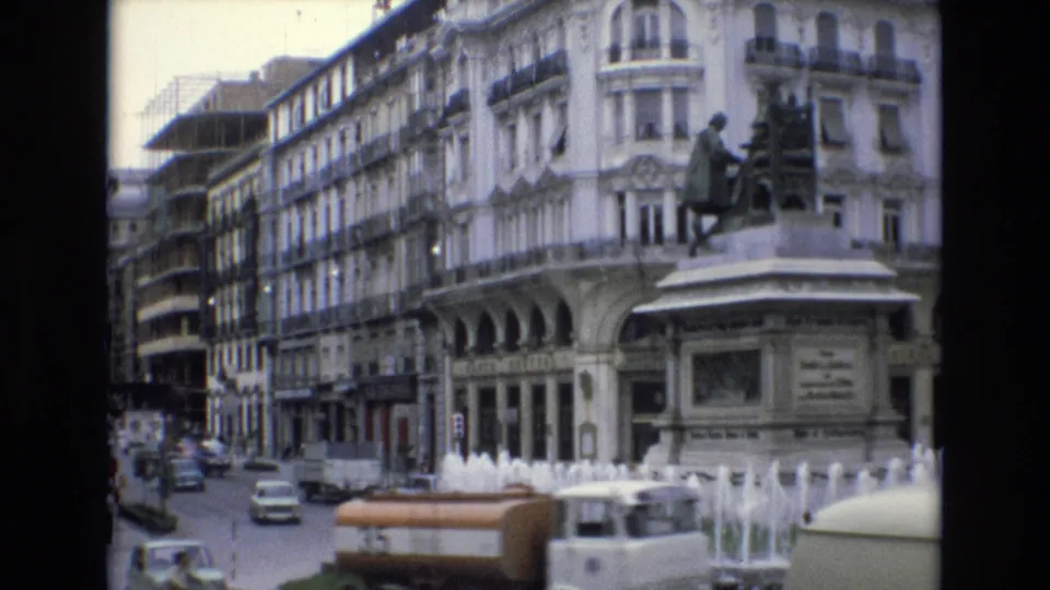 1972 Spain 8mm frame showing an orange-and-white vehicle near a spraying fountain, with period cars, pedestrians, a statue, and a large ornate building along the street.