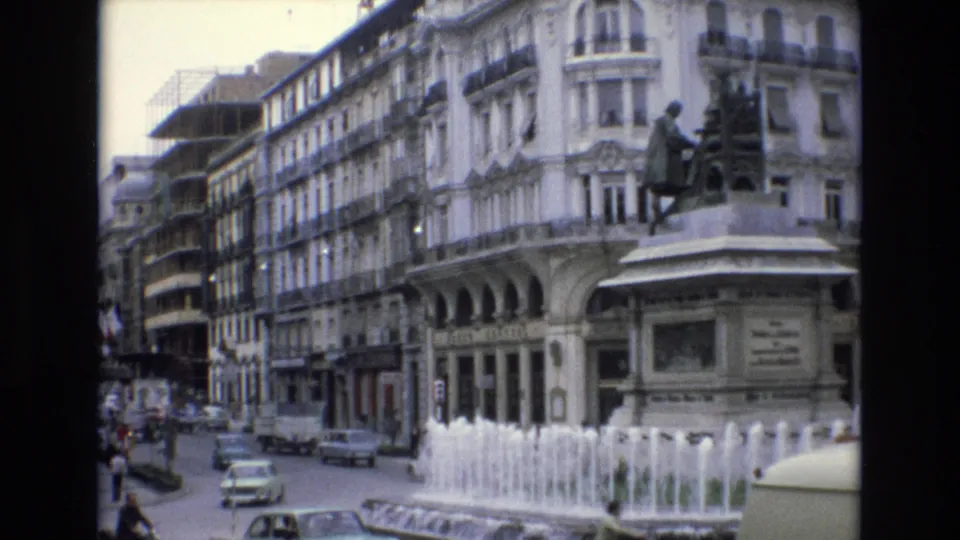 1972 Spain city street frame with fountain jets in the foreground, cars on the road, a statue on a pedestal, and a large white building with decorative balconies.