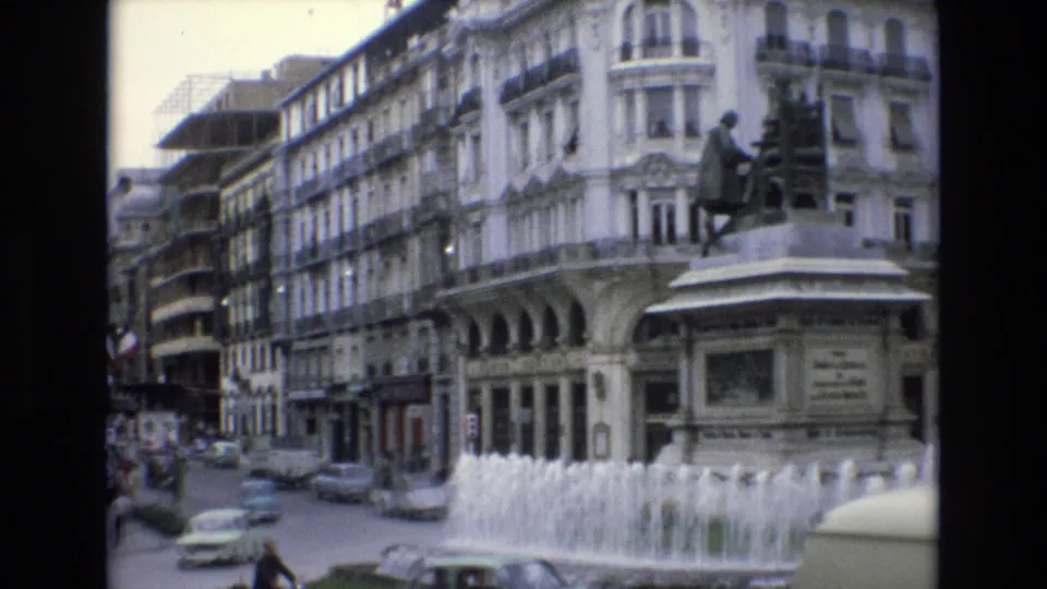Spain 1972 8mm frame showing a city square with a multi-jet fountain, a statue on a stone pedestal, classic cars, pedestrians, and multi-story buildings with balconies.
