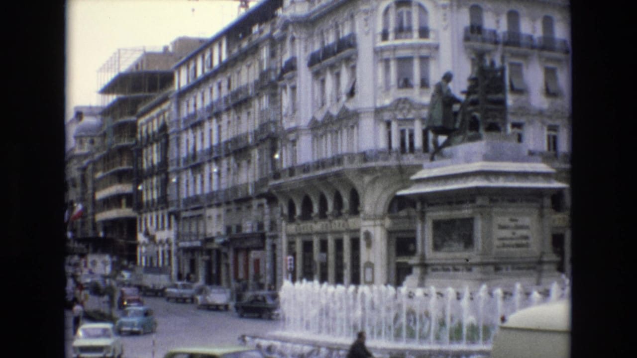 Spain 1972 8mm frame with cars moving around a traffic circle, a central fountain, a statue pedestal, pedestrians near the pavement, and ornate urban architecture behind them.