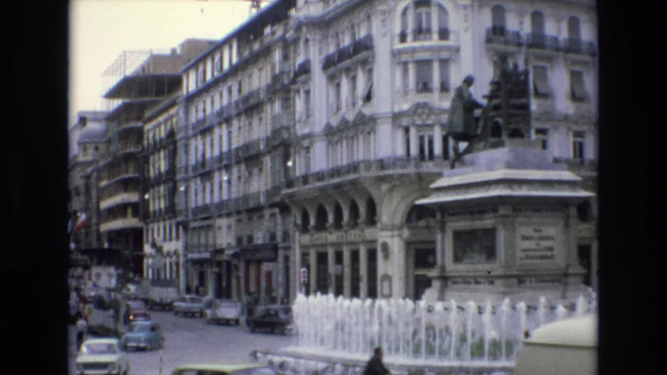 Spain 1972 8mm frame with cars moving around a traffic circle, a central fountain, a statue pedestal, pedestrians near the pavement, and ornate urban architecture behind them.