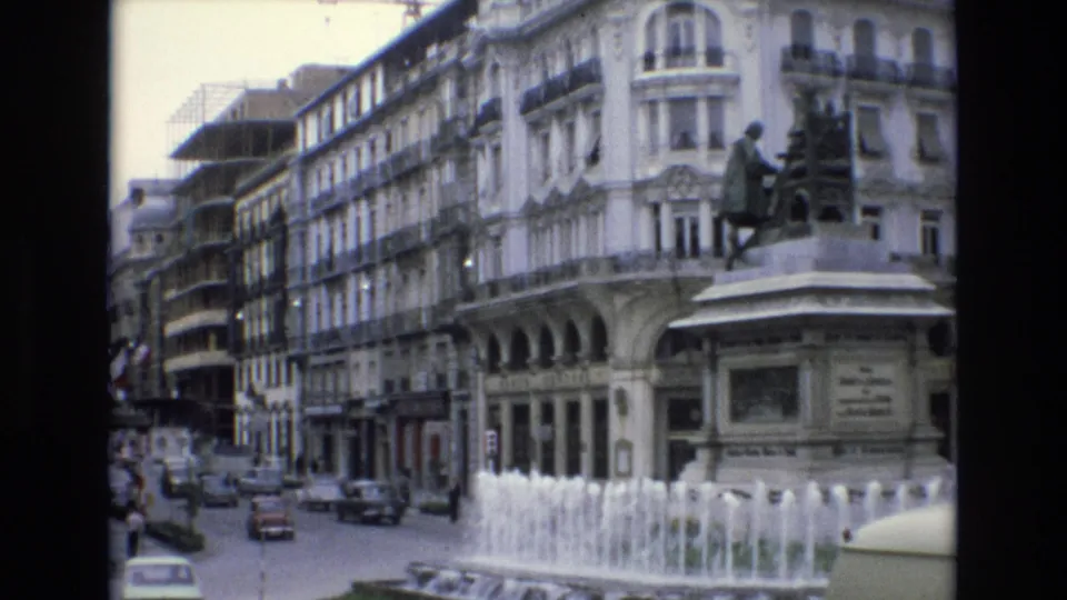 Spain 1972 8mm frame showing the fountain and seated statue more prominently, with traffic, pedestrians, storefronts, and tall buildings lining the square.