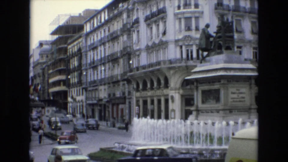 Spain 1972 8mm frame capturing the traffic circle from a wider angle, with the fountain in the foreground, statue pedestal at right, cars entering the square, pedestrians, and dense urban facades.