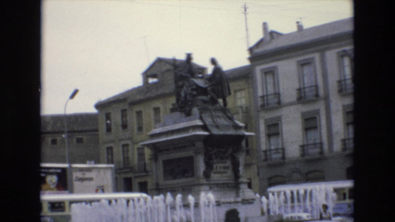 1972 Spain 8mm frame showing a bronze monument on a stone pedestal behind fountain jets, with balcony-fronted buildings and a streetlamp in the background.