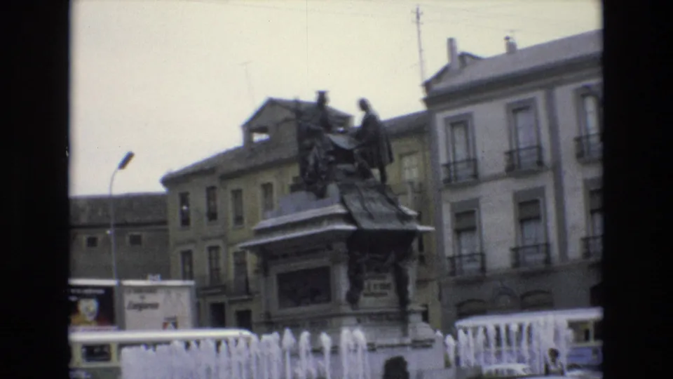 1972 Spain 8mm frame showing a bronze monument on a stone pedestal behind fountain jets, with balcony-fronted buildings and a streetlamp in the background.