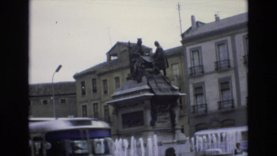 1972 Spain 8mm frame with a white-and-blue bus moving across the foreground, partly blocking the fountain and bronze monument in a city square.