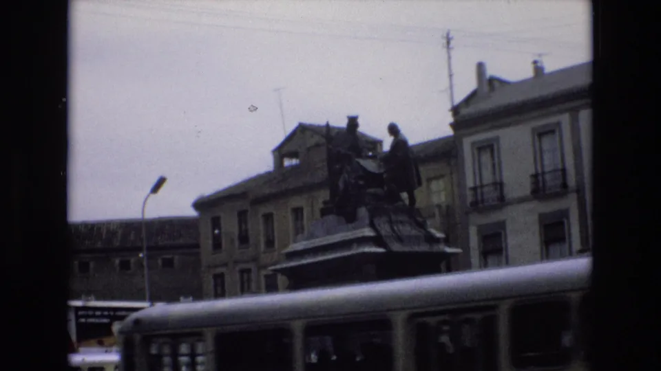 1972 Spain 8mm frame where a passing bus obscures much of the fountain while the bronze monument, streetlamp, and older buildings remain visible above it.