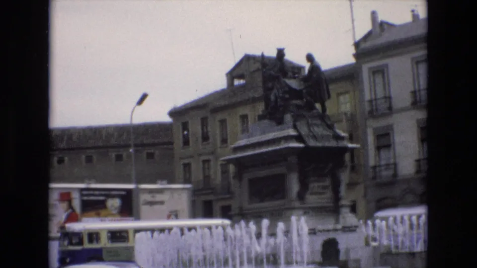 1972 Spain 8mm frame showing fountain jets in the foreground, a bronze monument on a stone base, surrounding buildings, and a bus or traffic element near the left edge.