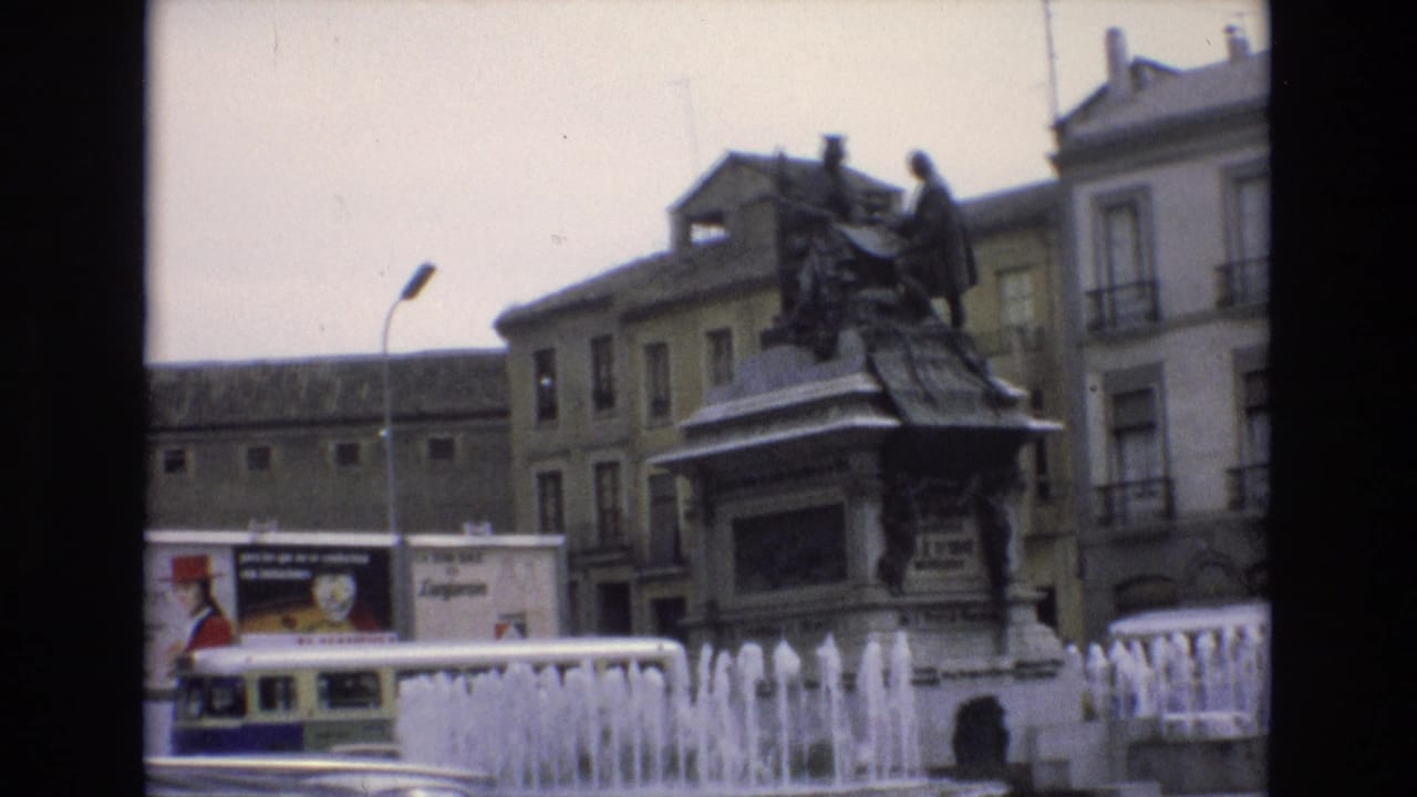 1972 Spain home-movie frame showing a monument statue above bubbling fountain jets, with multi-story buildings, a streetlamp, advertising panels, and a passing bus or coach.