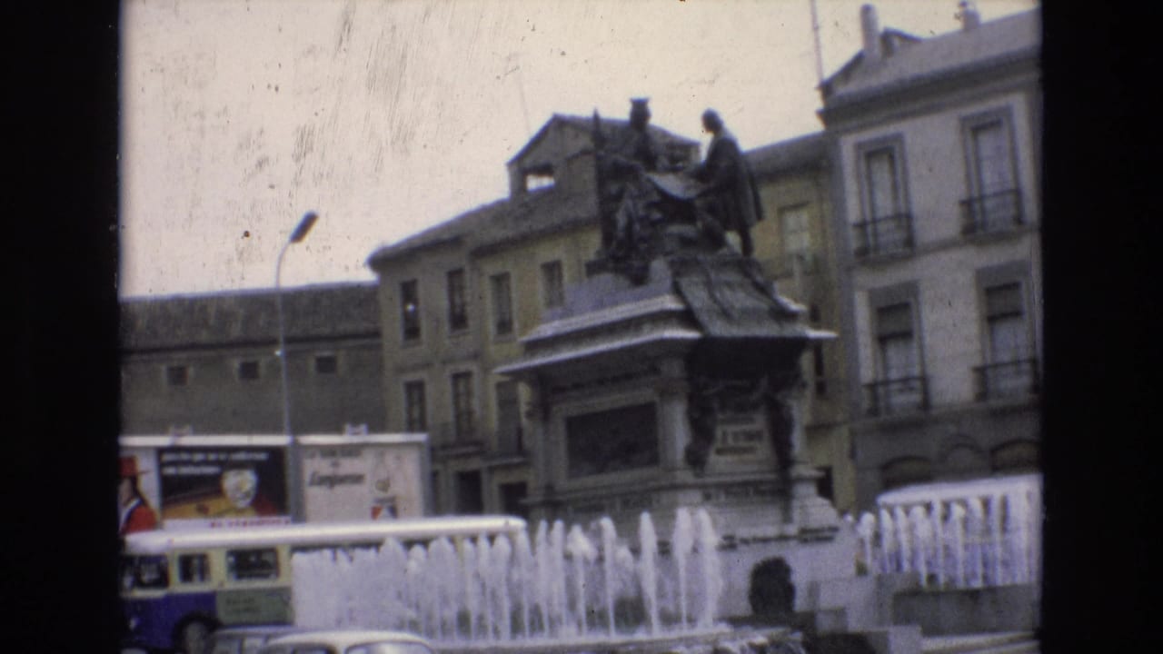 Statue and fountain jets in front of older buildings with a bus moving along the lower edge.