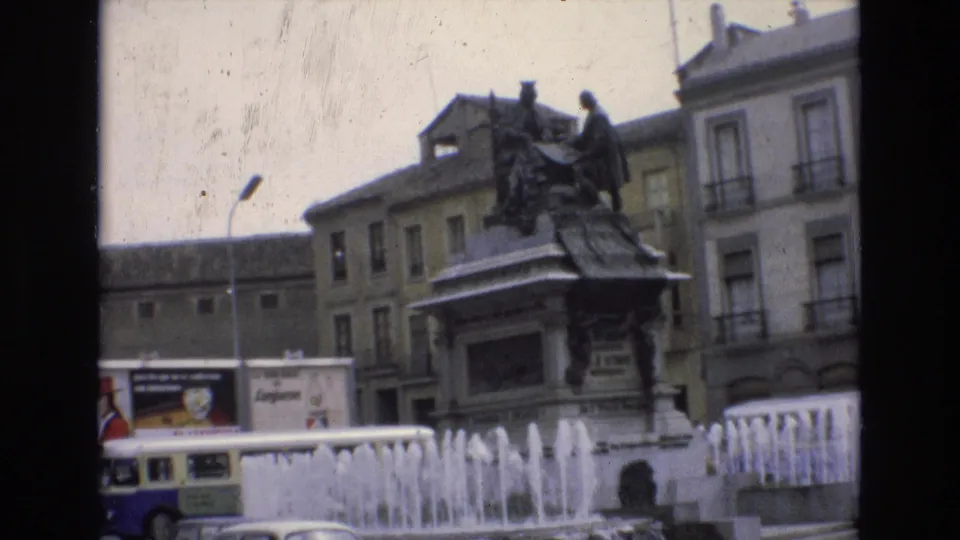 1972 Spain frame of a public square monument and active fountain, with older buildings behind it and a partially visible bus or coach crossing the foreground.
