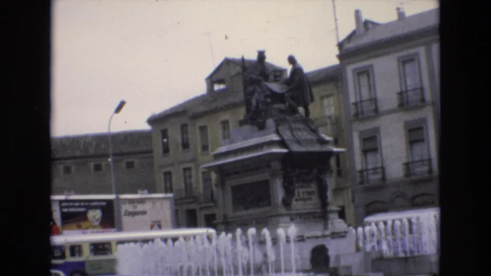 Slightly tilted 1972 Spain home-movie view of the statue and fountain jets, with urban building facades, a curved streetlamp, and a bus partly visible along the lower left.