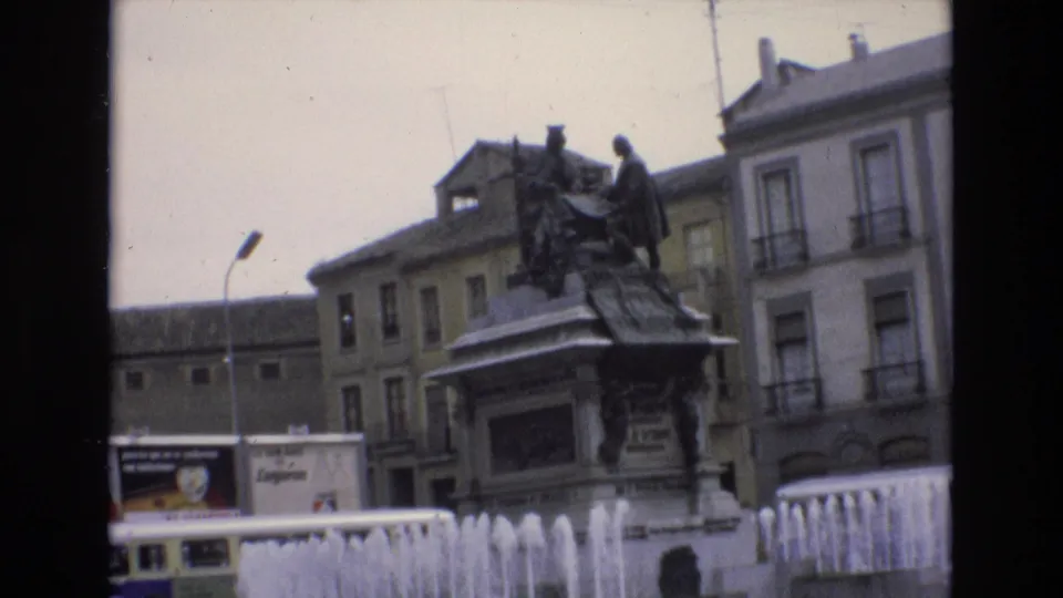 1972 Spain 8mm frame holding on the civic statue and bubbling fountain, with surrounding buildings, advertising panels, and dark contact-sheet borders visible.