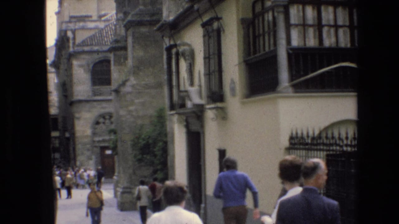 In Spain in 1972, pedestrians in period clothing walk along a narrow street lined with historic stone facades and balconies.