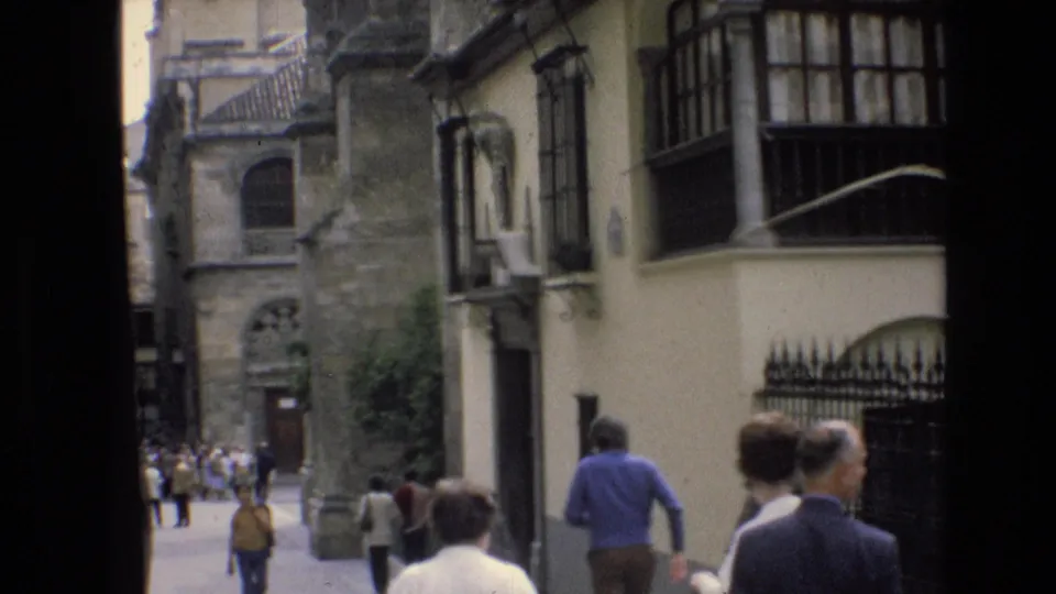 In Spain in 1972, pedestrians in period clothing walk along a narrow street lined with historic stone facades and balconies.