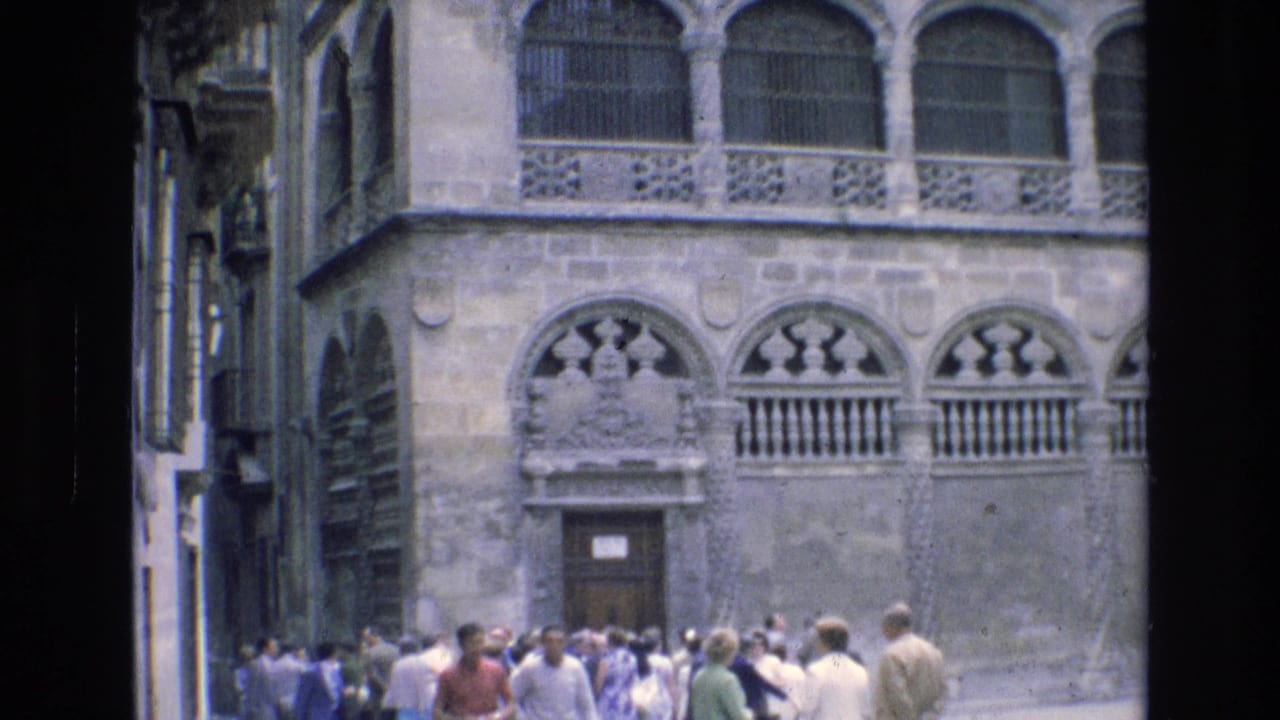 Visitors gather near an ornate historic stone building in Spain, with arched windows, balcony railings, and carved facade details visible.