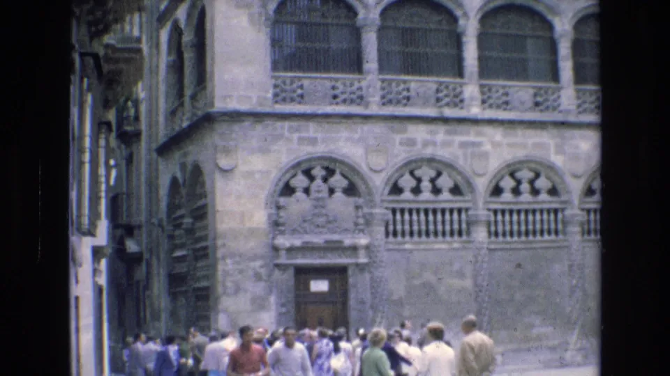 Visitors gather near an ornate historic stone building in Spain, with arched windows, balcony railings, and carved facade details visible.