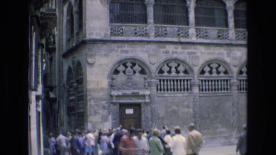 A crowd of pedestrians and visitors stands near a carved historic facade in Spain, with arched openings and decorative stonework filling the frame.