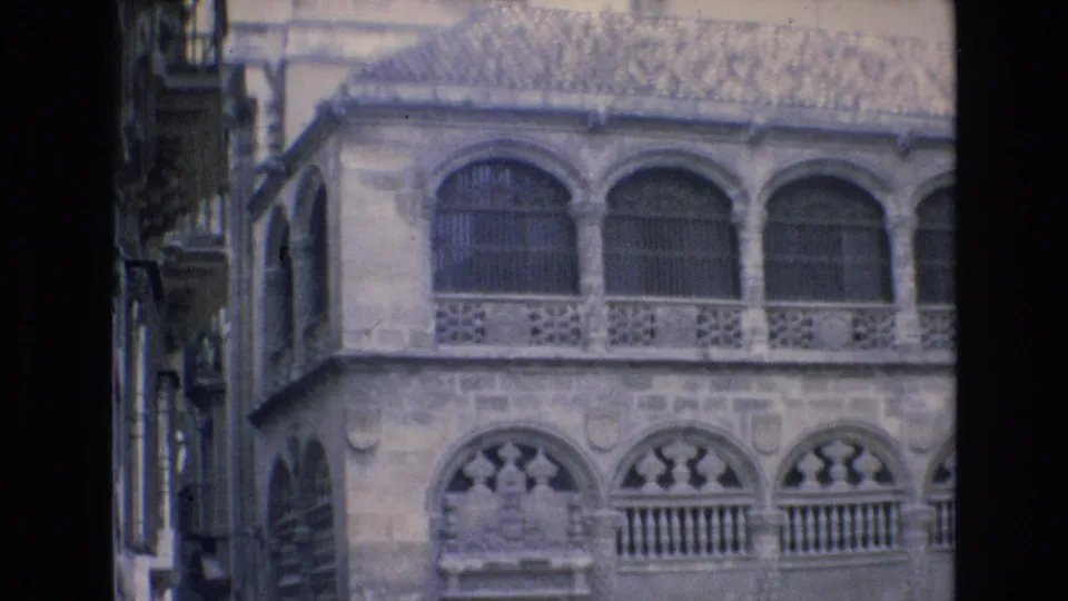 The frame centers on a historic stone building in Spain, showing arched upper windows, decorative railings, carved lower arches, and pedestrians below.