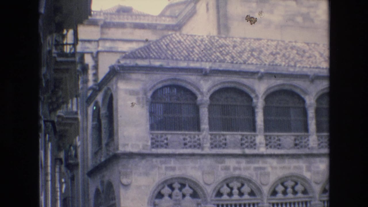 1972 Spain 8mm frame showing a historic stone facade with arched windows, decorative railing, tiled roof and shadowed framing from a narrow street view.