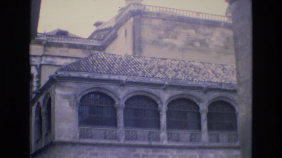 1972 Spain home-movie frame showing a historic stone exterior with repeated arched windows, decorative balcony railings, tiled roof and softly faded 8mm color.