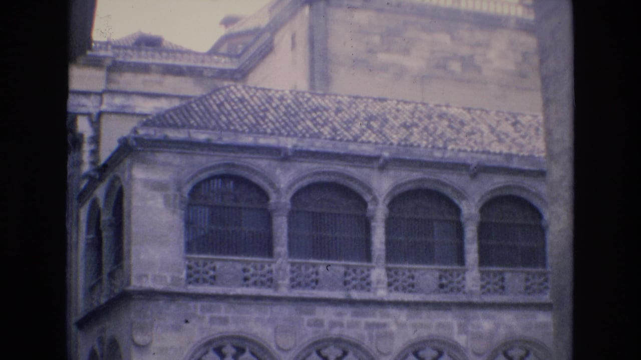 1972 Spain 8mm frame showing the upper facade of a historic stone building with arched windows, decorative railing, and a tiled roof edge.