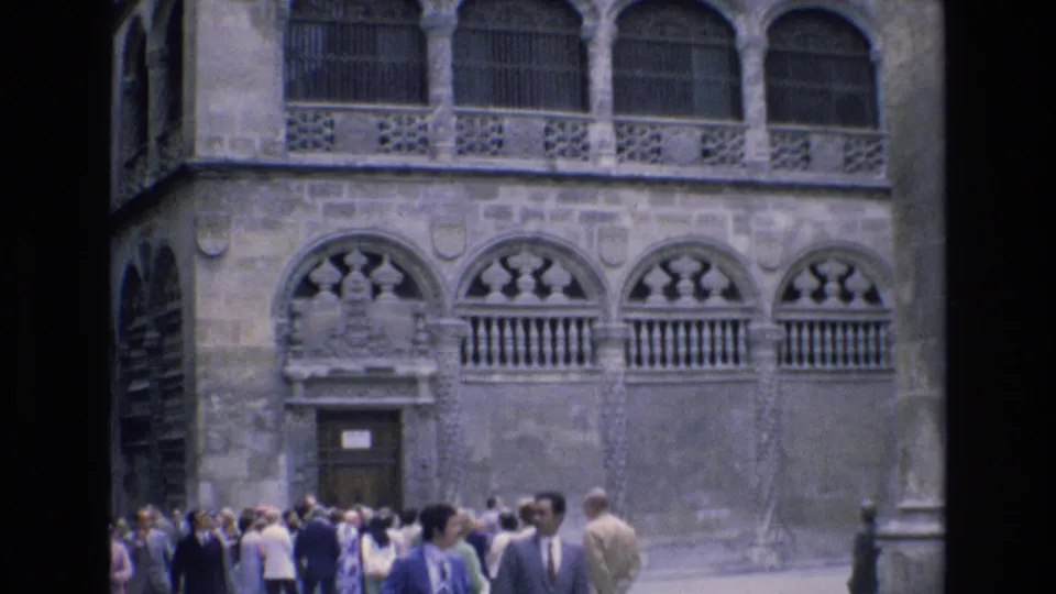 A 1972 Spain home-movie frame shows pedestrians in suits, dresses, and light clothing gathered near the entrance below the ornate stone facade.
