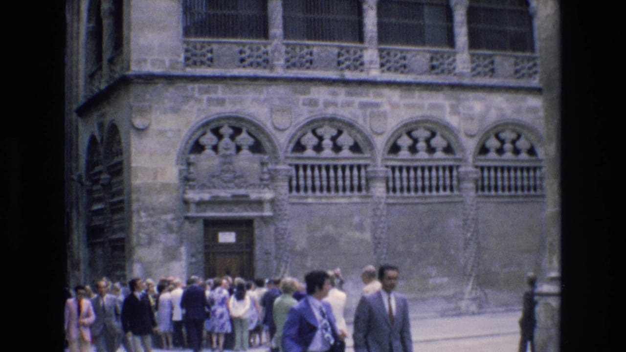 1972 Spain home-movie frame showing tourists in period clothing gathered near a historic stone facade with carved arched balcony details.