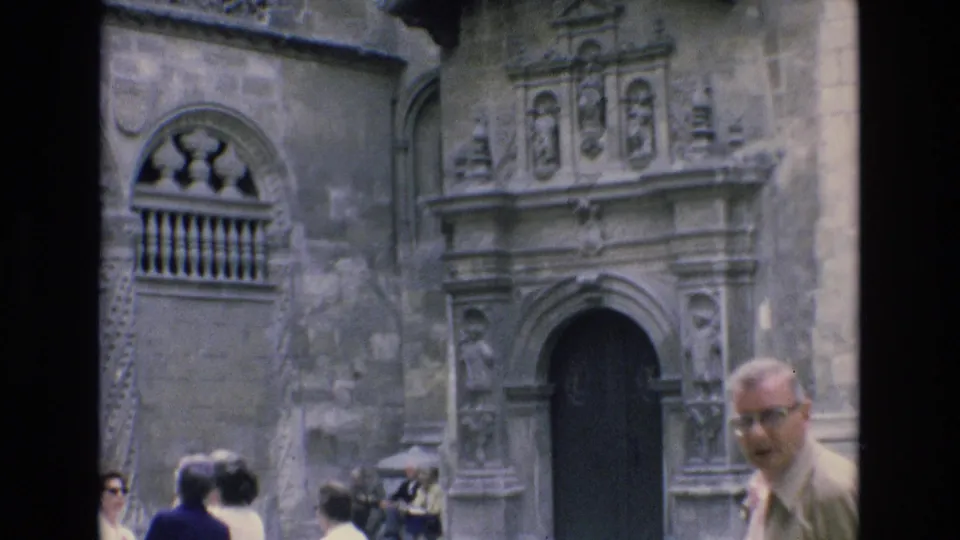 1972 Spain home-movie frame showing a man near the foreground and other tourists beside an ornate stone doorway on a historic facade.