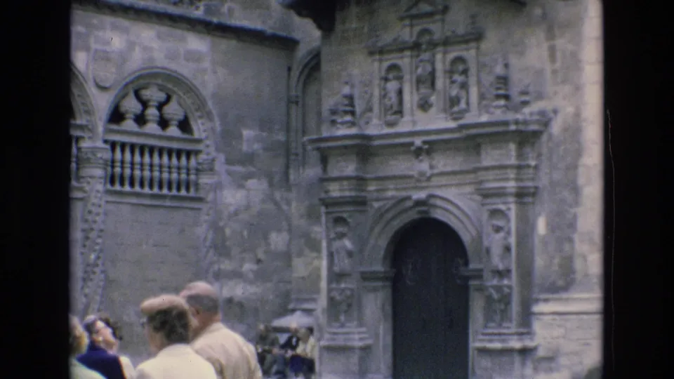 1972 Spain frame showing tourists gathered at street level in front of a historic stone facade with carved doorway and arched balcony opening.