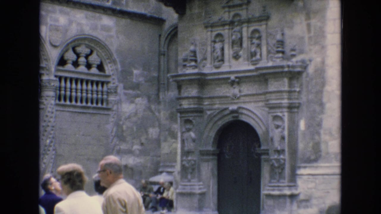 1972 Spain 8mm frame showing tourists in the foreground before a weathered stone building facade with a carved arched entrance and balcony detail.