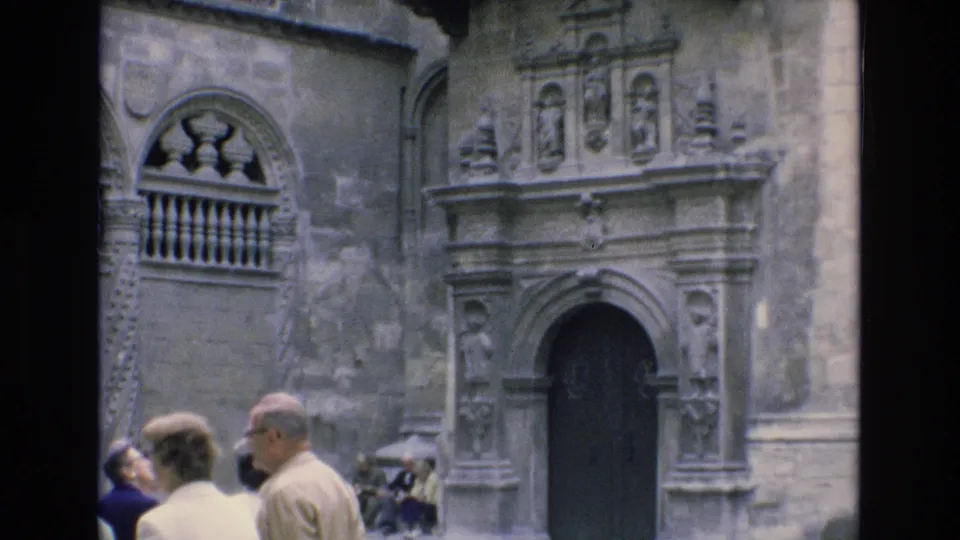 1972 Spain 8mm frame showing tourists in the foreground before a weathered stone building facade with a carved arched entrance and balcony detail.
