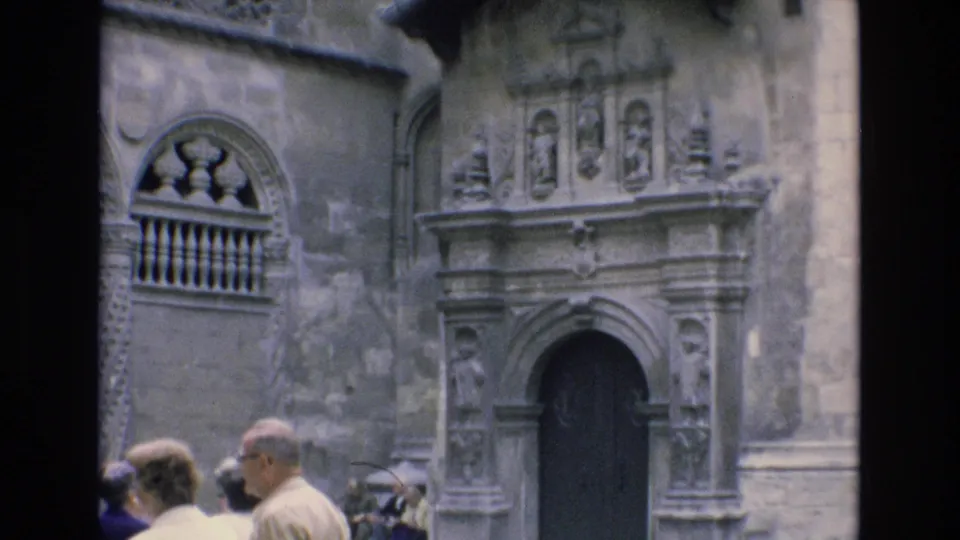 1972 Spain travel footage frame with visitors partly visible below a carved stone doorway, sculpted facade elements, and an arched balcony opening.