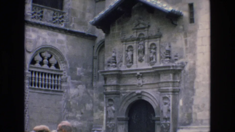 1972 Spain 8mm frame focused on the weathered stone facade, showing the arched entrance, sculptural doorway surround, balcony opening, and partial tourist figures.