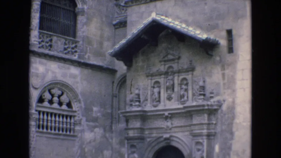 1972 Spain home-movie frame showing the upper part of an ornate stone facade with carved decorative panels, balcony openings, weathered wall surface, and tiled roof edge.
