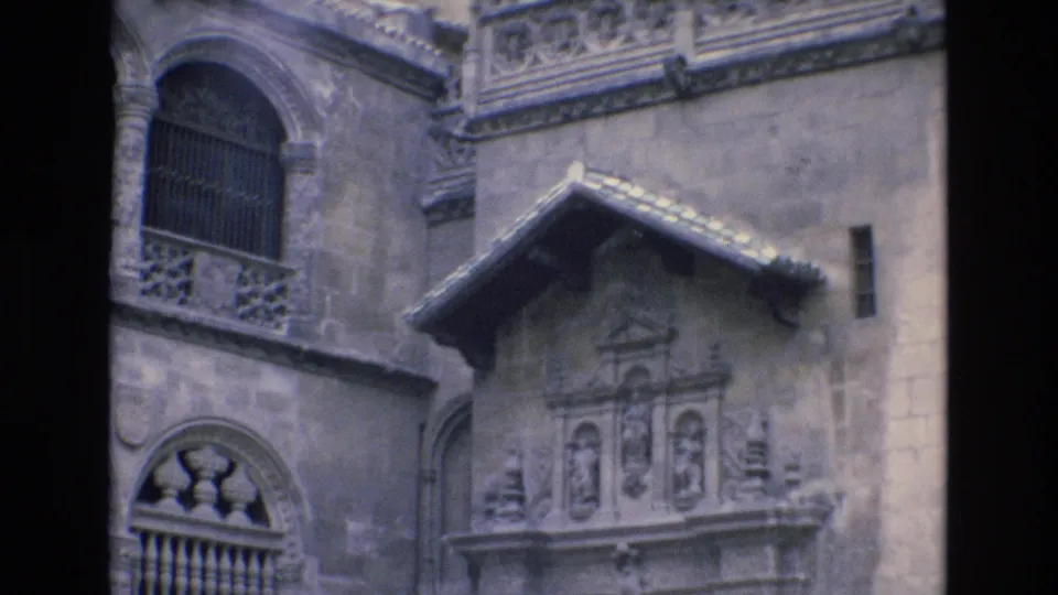 1972 Spain 8mm frame of a historic facade detail with a barred arched window, relief carving beneath a tiled gable, and decorative stone railing above.