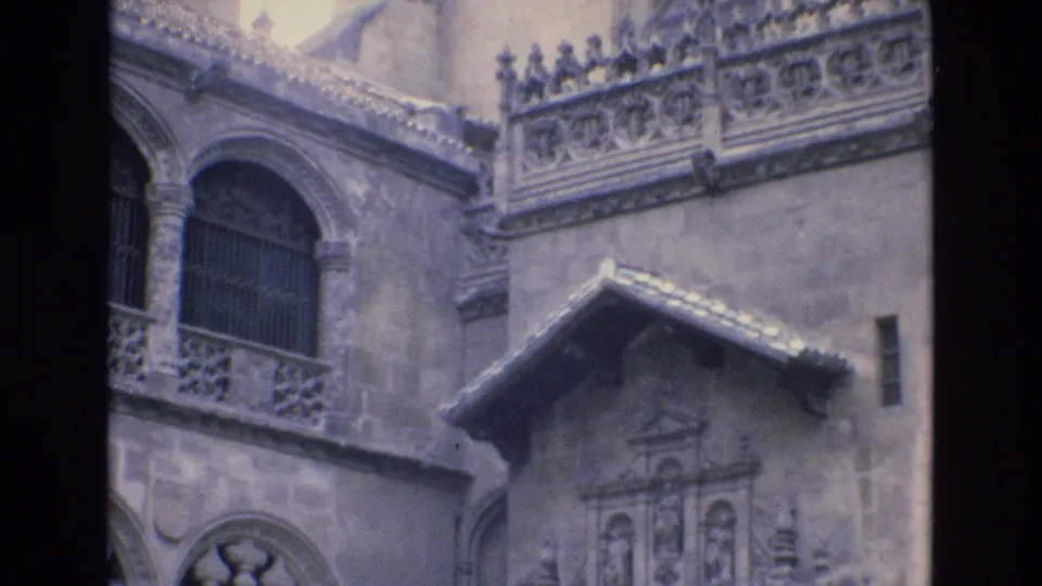 1972 Spain 8mm frame showing a closer visitor view of arched stone windows, ornamental balcony railings, carved facade panels, and roof tile edges.